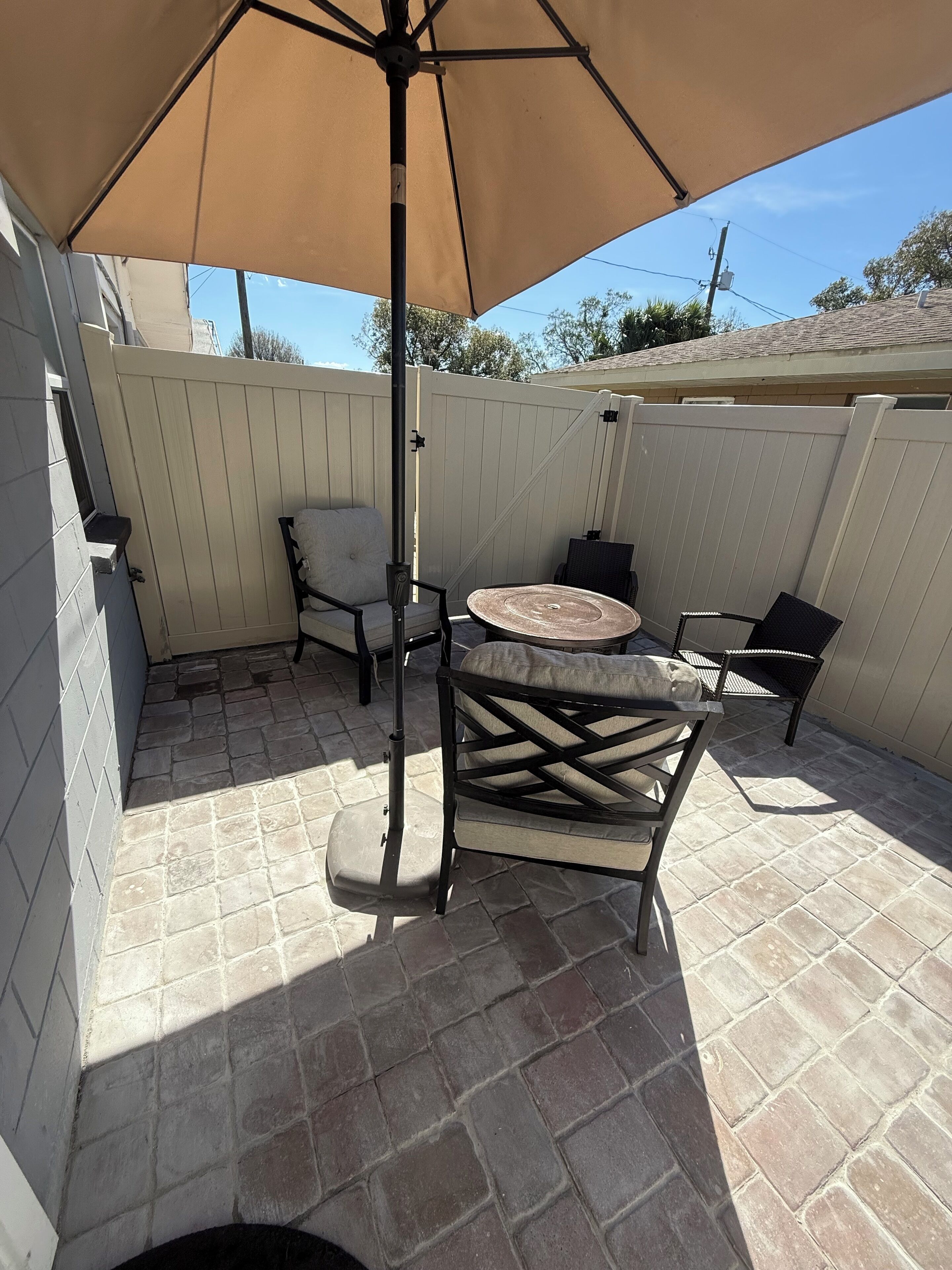 A patio area with a table and chairs under a large umbrella, surrounded by a white fence and a stone-tiled floor.