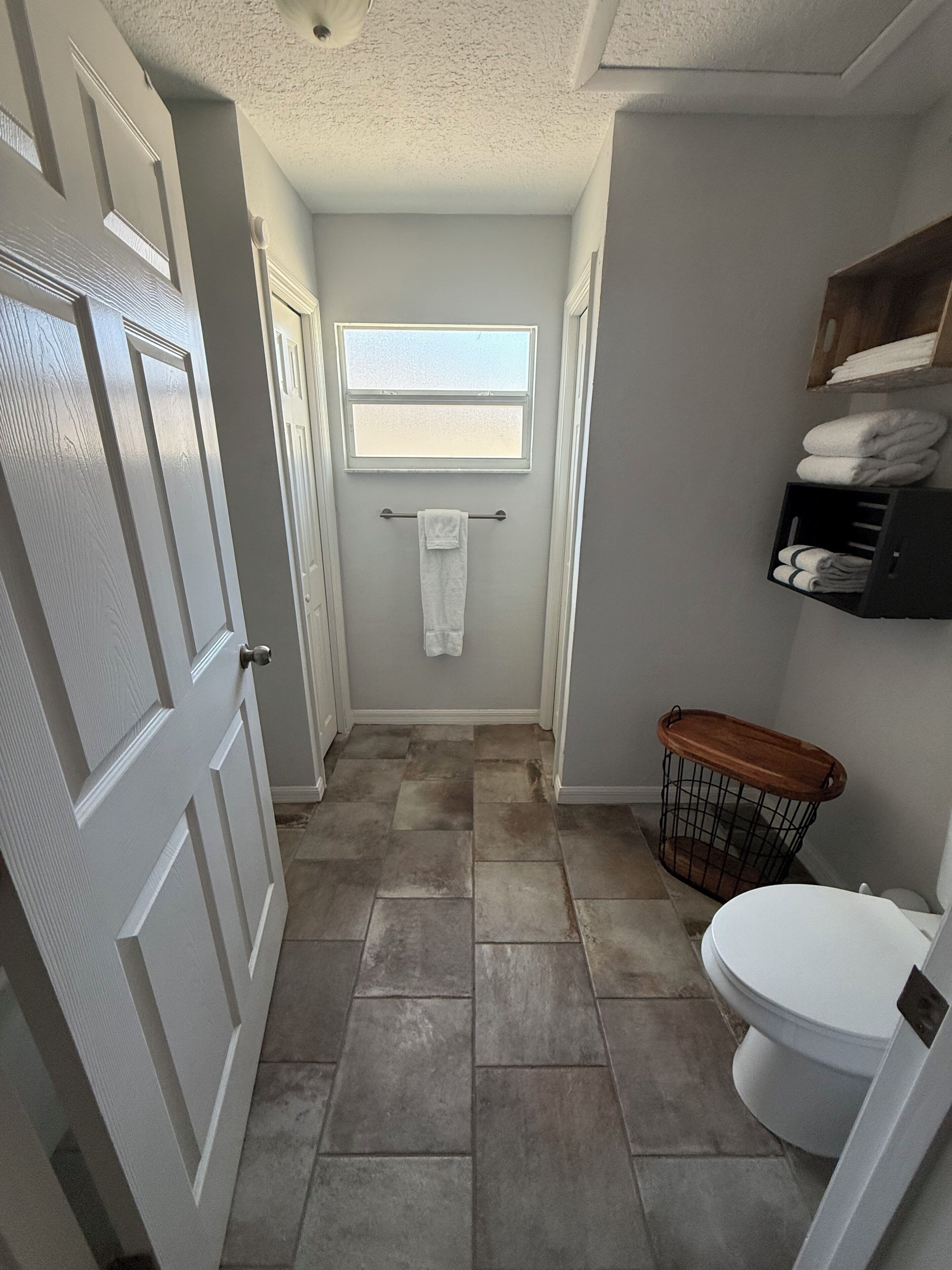 A narrow bathroom with a white toilet, a window, a towel rack, and a basket.