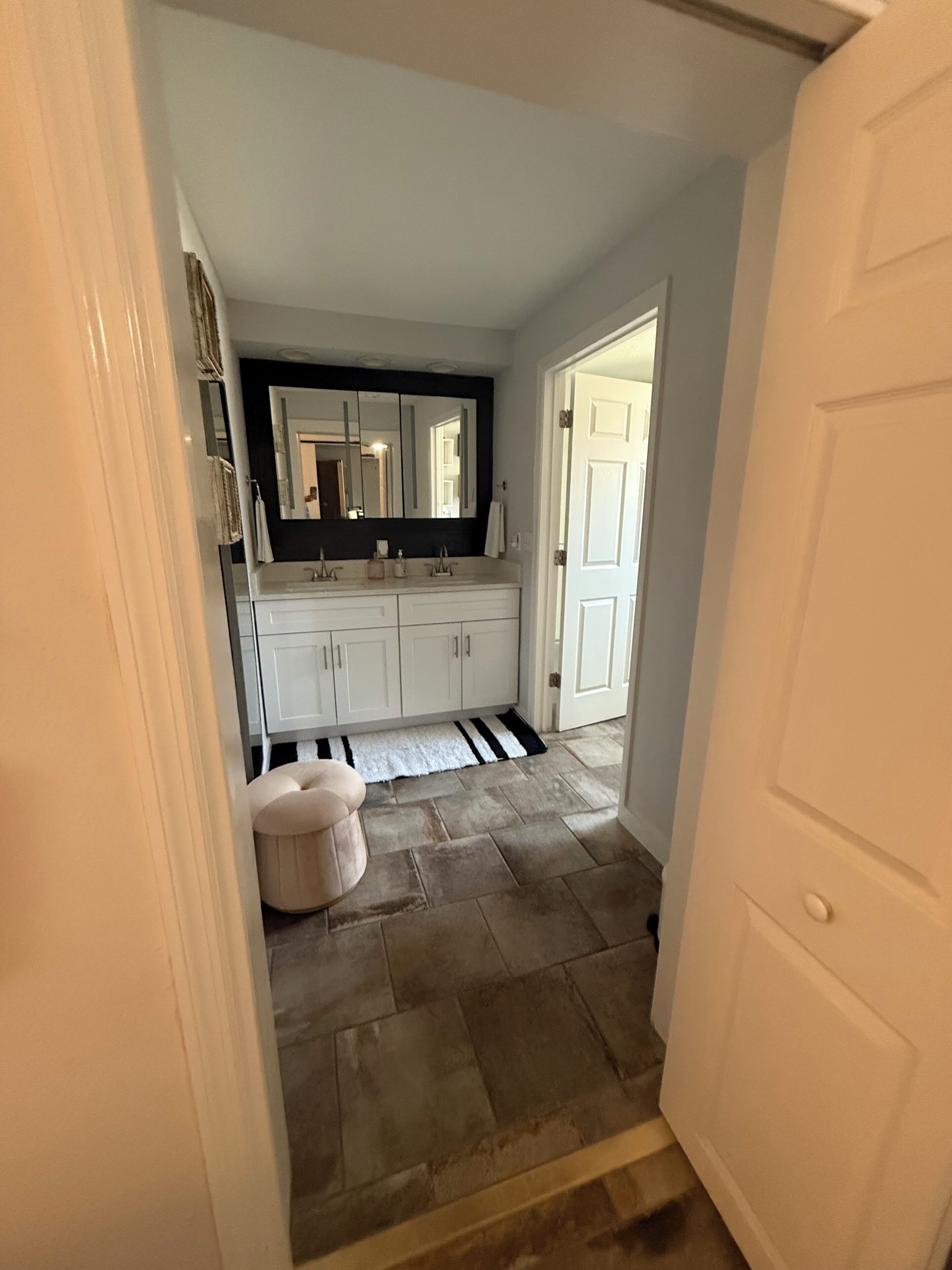 A hallway leading to a bathroom with white cabinets, a black countertop, and a large mirror.