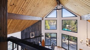 Un interior moderno con techo de madera inclinado, ventanas amplias que ofrecen vistas a un lago y un bosque, y una tabla de surf sobre una estantería.