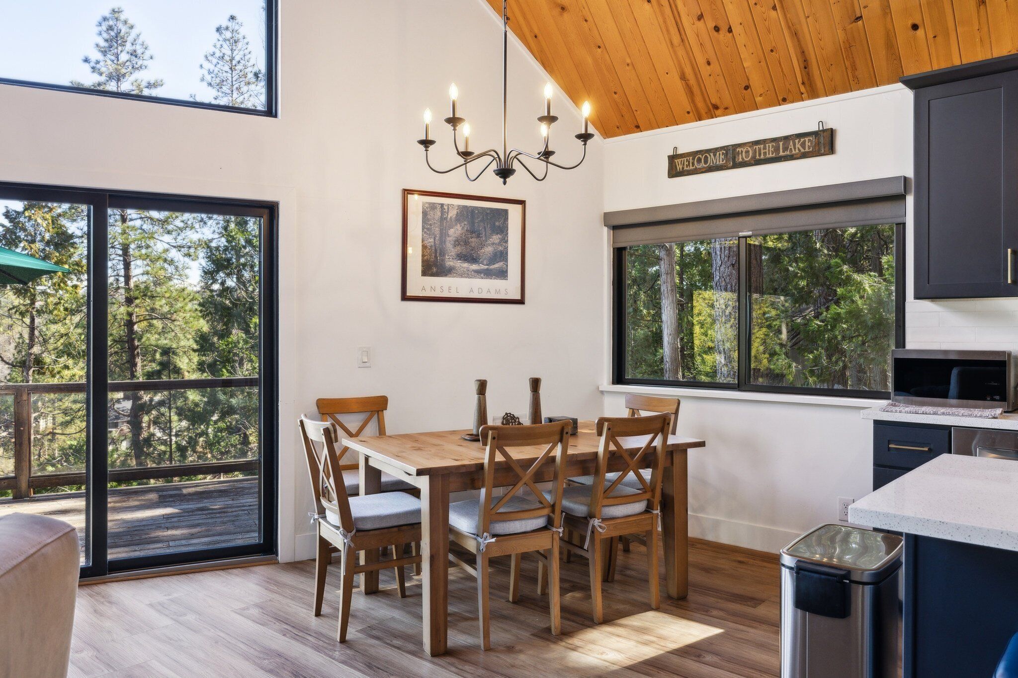 Un comedor con mesa y sillas de madera, una araña y vistas a árboles a través de amplios ventanales.