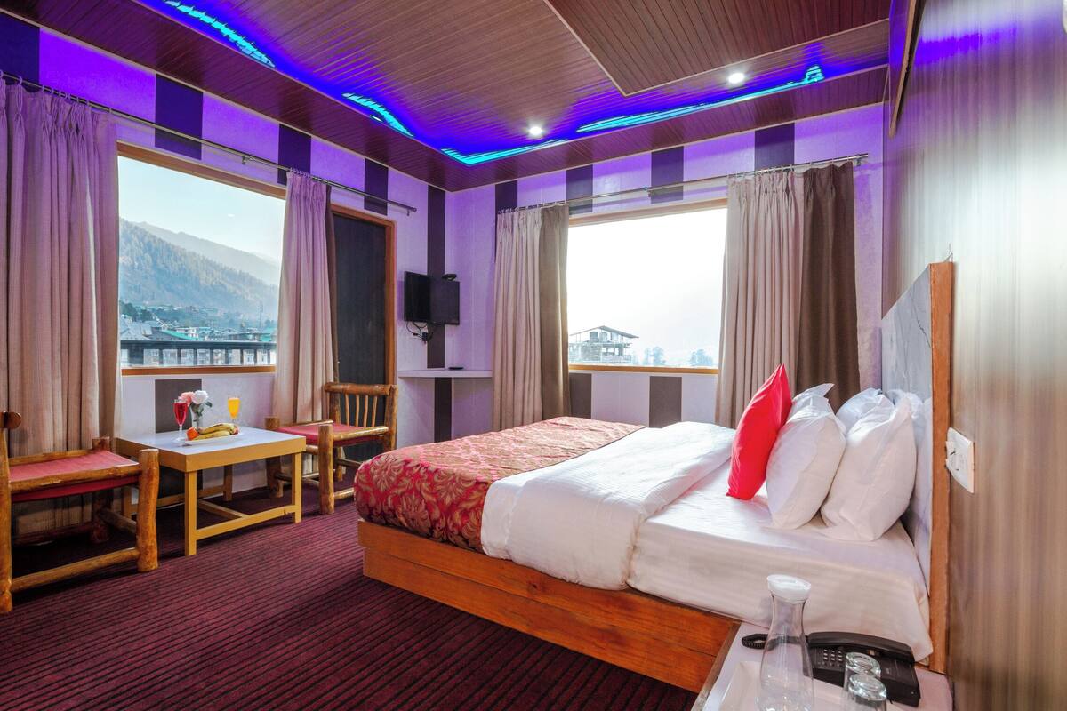 A hotel room with a bed, a table with a chair, a small table with a bottle and glasses, and a view of a mountain and a bridge.