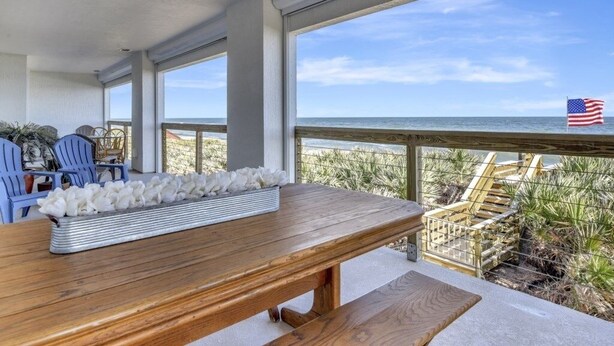 Una mesa de madera con un arreglo floral blanco, sillas azules y vista a la playa con una bandera estadounidense.