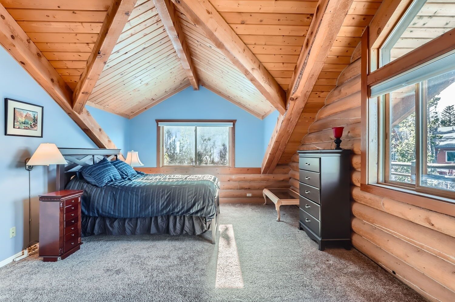 Une chambre avec un plafond en pente, un lit, une commode, une table de chevet, une lampe, une fenêtre et un tableau accroché au mur.