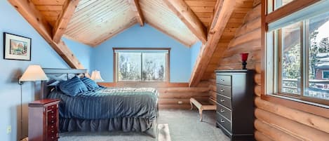 Une chambre avec un plafond en pente, un lit, une commode, une table de chevet, une lampe, une fenêtre et un tableau accroché au mur.