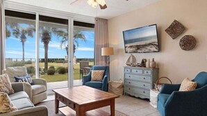 A living room with a view of palm trees and the ocean, a ceiling fan, a wooden coffee table, and a blue armchair. - Beach Club Bristol 101 3 Bedroom Condo (Gulf Shores)
