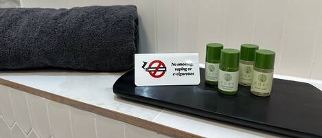 Letrero que prohíbe fumar, vapear o cigarrillos electrónicos, y tres botellas de gel de baño Eden sobre una bandeja negra.