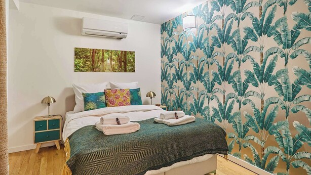 Un dormitorio con cama, mesita de noche, aire acondicionado instalado en la pared, un cuadro y papel tapiz con hojas de plátano.