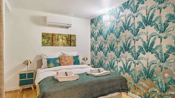Une chambre à coucher comprenant un lit, une table de chevet, un climatiseur fixé au mur, un tableau et un papier peint à motifs de feuilles de bananier.