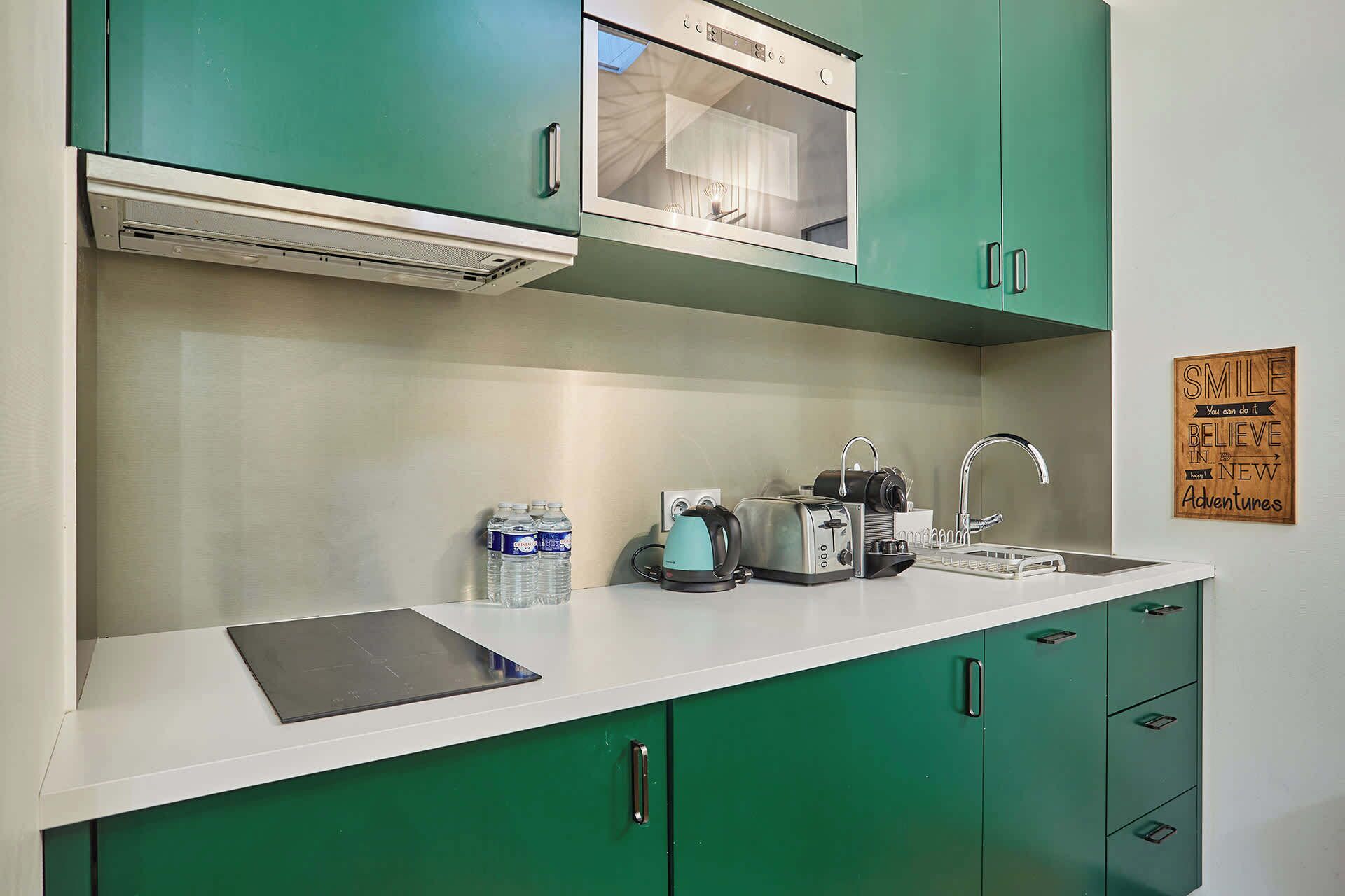 Une cuisine avec des meubles de couleur verte, un plan de travail blanc, un grille-pain, une bouilloire et un évier.