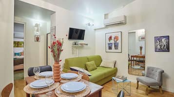 Un coin repas avec une table en bois dressée pour quatre personnes, un canapé vert, un fauteuil gris et un téléviseur fixé au mur.