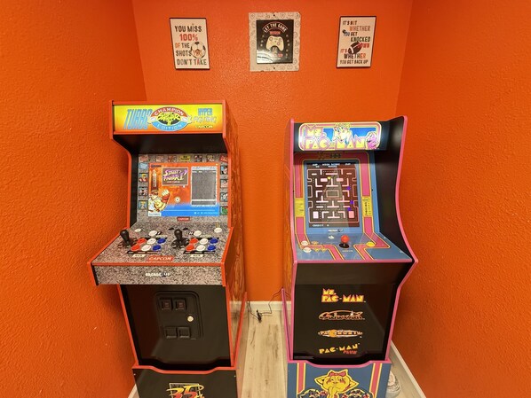 Dos máquinas de videojuegos de arcade, una para 'Street Fighter II' y otra para 'Pac-Man', frente a una pared naranja decorada con carteles de motivación.