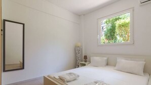 A bedroom with a bed, a bedside table with a lamp, a window with a view of greenery, and a mirror on the wall. - Dreamy Cove, Artistic Hideaway near Floisvos 1BD (Paleo Faliro)