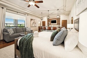 A modern hotel room with a large bed, a sofa, a dining area, and a view of the outdoors.