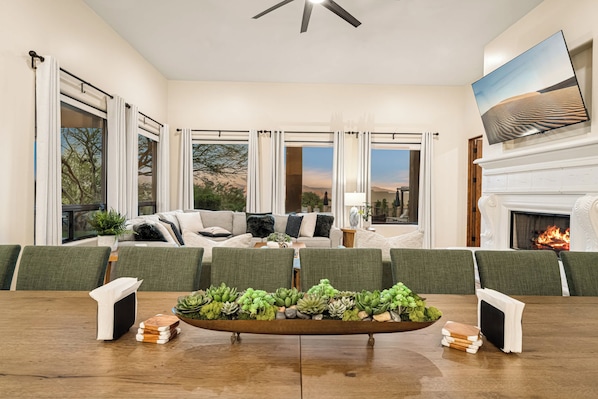 A spacious living room with a fireplace, a large window with curtains, a dining table with chairs, and a sofa with cushions.