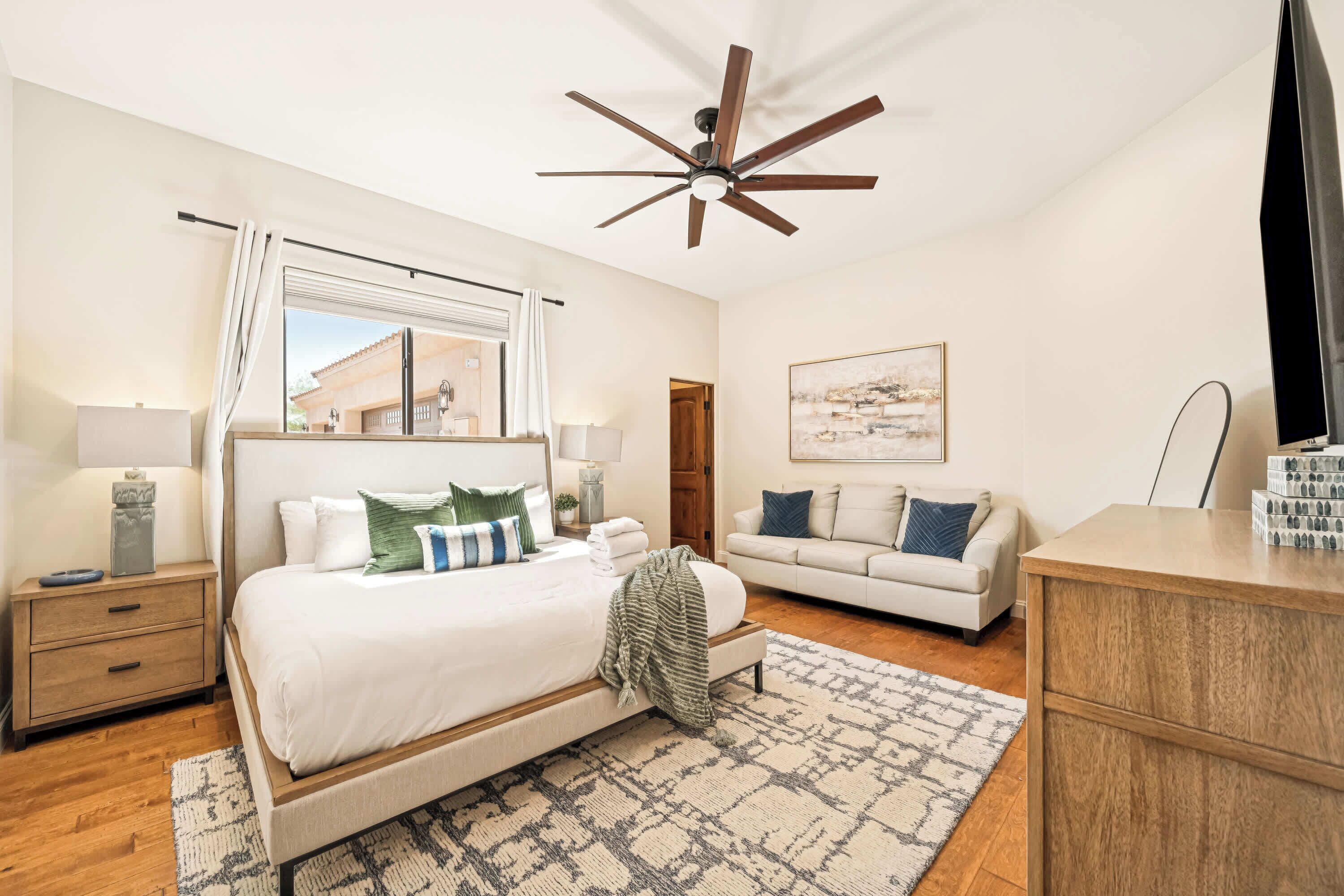 A bedroom with a bed, a nightstand, a dresser, a ceiling fan, a window with curtains, a wall-mounted TV, and a painting.
