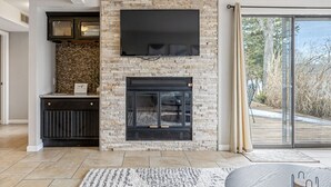 Un salon moderne avec une cheminée en pierre, un téléviseur à écran plat fixé au-dessus, et une porte-fenêtre coulissante donnant sur une terrasse extérieure.