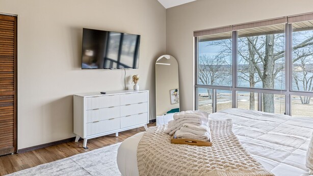 Une chambre moderne avec un grand lit, un téléviseur fixé au mur et une vue sur l’extérieur grâce à de grandes fenêtres.