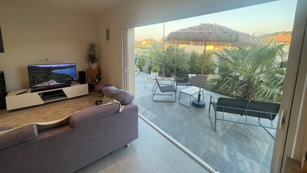 Una sala de estar moderna con un televisor de pantalla plana, un sofá y un balcón con mobiliario de exterior y plantas.