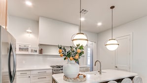 A modern kitchen with white cabinetry, a central island with chairs, a large vase with oranges, and pendant lighting. - Carlton Landing-Shingle Ready to Mingle (Carlton Landing)