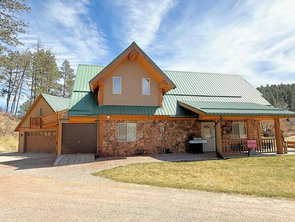 Un chalet à deux étages avec une façade en pierre, un toit en métal vert et une terrasse en bois.