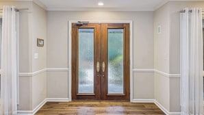 Un pasillo con una puerta doble de madera con paneles de vidrio, paredes blancas y suelo de madera.