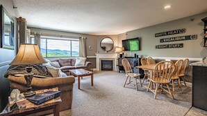 A cozy living room with a fireplace, a dining area with wooden chairs, a TV mounted on the wall, and a view of the outdoors through large windows. - Two bedroom North Peak Condo (Newry)
