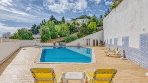 A swimming pool with yellow and white lounge chairs, a tiled patio, and a view of a hillside with trees and buildings.