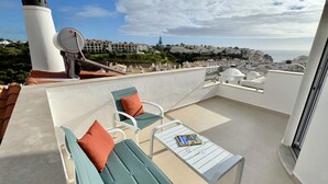 A rooftop terrace with two green sofas, a small table, and a white chair. A white satellite dish is visible on the left. The terrace overlooks a coastal town with buildings and a lighthouse in the background.