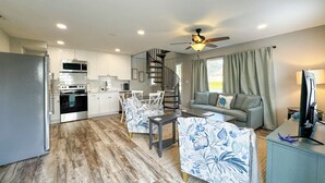 A modern kitchen with white cabinets, a microwave, and a stove. There is a dining area with a table and chairs, and a living room with a sofa and a TV.