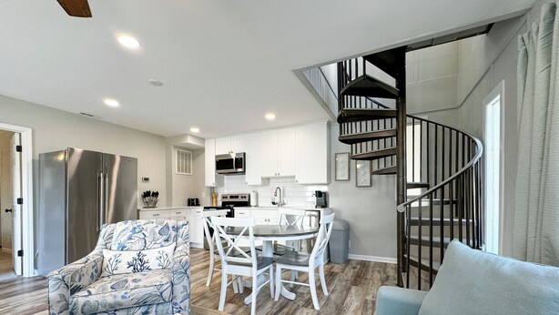 A modern living room with a sofa, a coffee table, and a patterned armchair. There is a dining area with a table and chairs, and a kitchen with white cabinets and a black staircase.