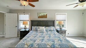 A bedroom with a large bed, two nightstands, a ceiling fan, and two windows with lamps.