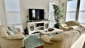 Een woonkamer met een televisie op een witte kast, beige banken, een salontafel en een grote plant.