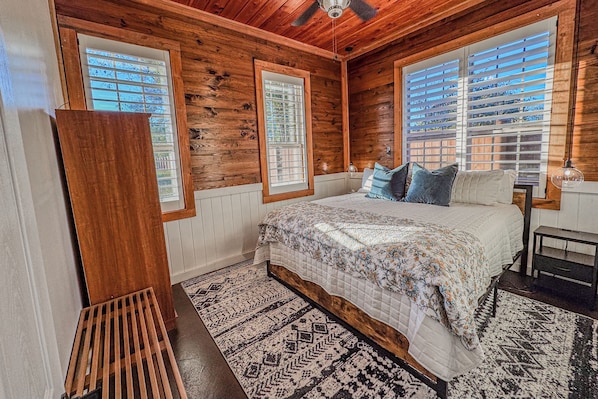 Un dormitorio con techo de madera, una cama con ropa de cama floral, una mesita de noche y una ventana con persianas.