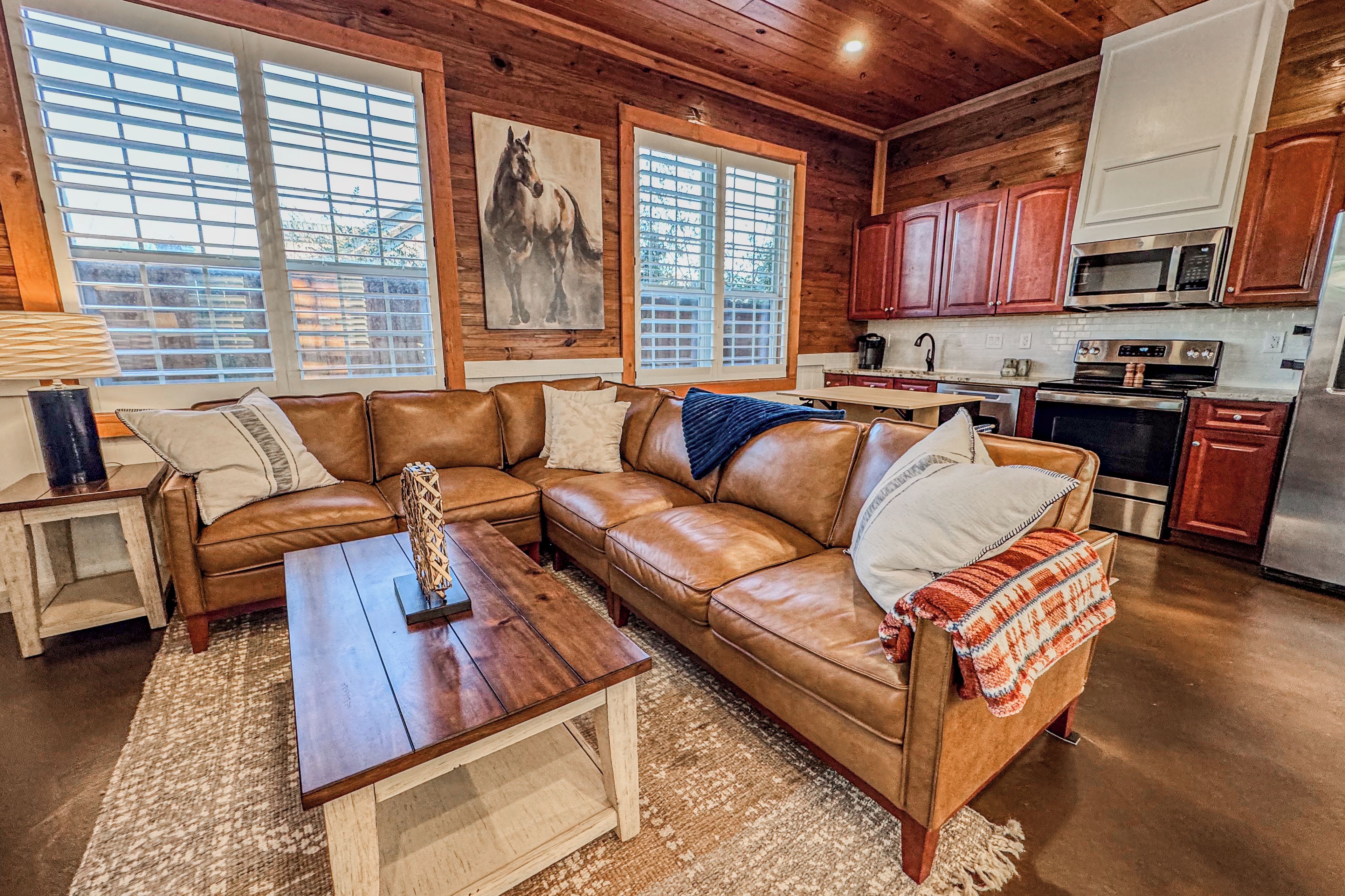 A cozy living room with a brown leather sectional sofa, a wooden coffee table, and a large window with blinds.