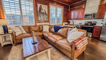 A cozy living room with a brown leather sectional sofa, a wooden coffee table, and a large window with blinds.