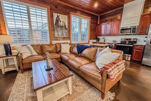 A cozy living room with a brown leather sectional sofa, a wooden coffee table, and a large window with blinds.