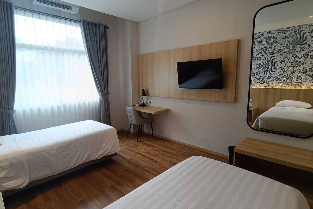 A hotel room with two single beds, a desk with a chair, a flat-screen TV mounted on the wall, and a large mirror.