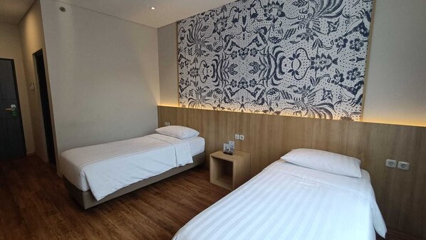 A hotel room with two single beds, a wooden headboard, a nightstand with a lamp, and a patterned wall hanging. - Adia Suites Hotel (South Tangerang)