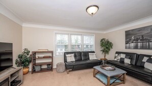 A living room with a black leather sofa, a grey ottoman, a wooden coffee table, a TV stand, and a large window with blinds. - Kenwood Keys 2 – Stay for 6! (Cincinnati)