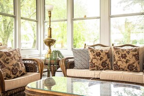Ein gemütliches Wohnzimmer mit Korbmöbeln, einem Glastisch und großen Fenstern mit Blick auf viel Grün.