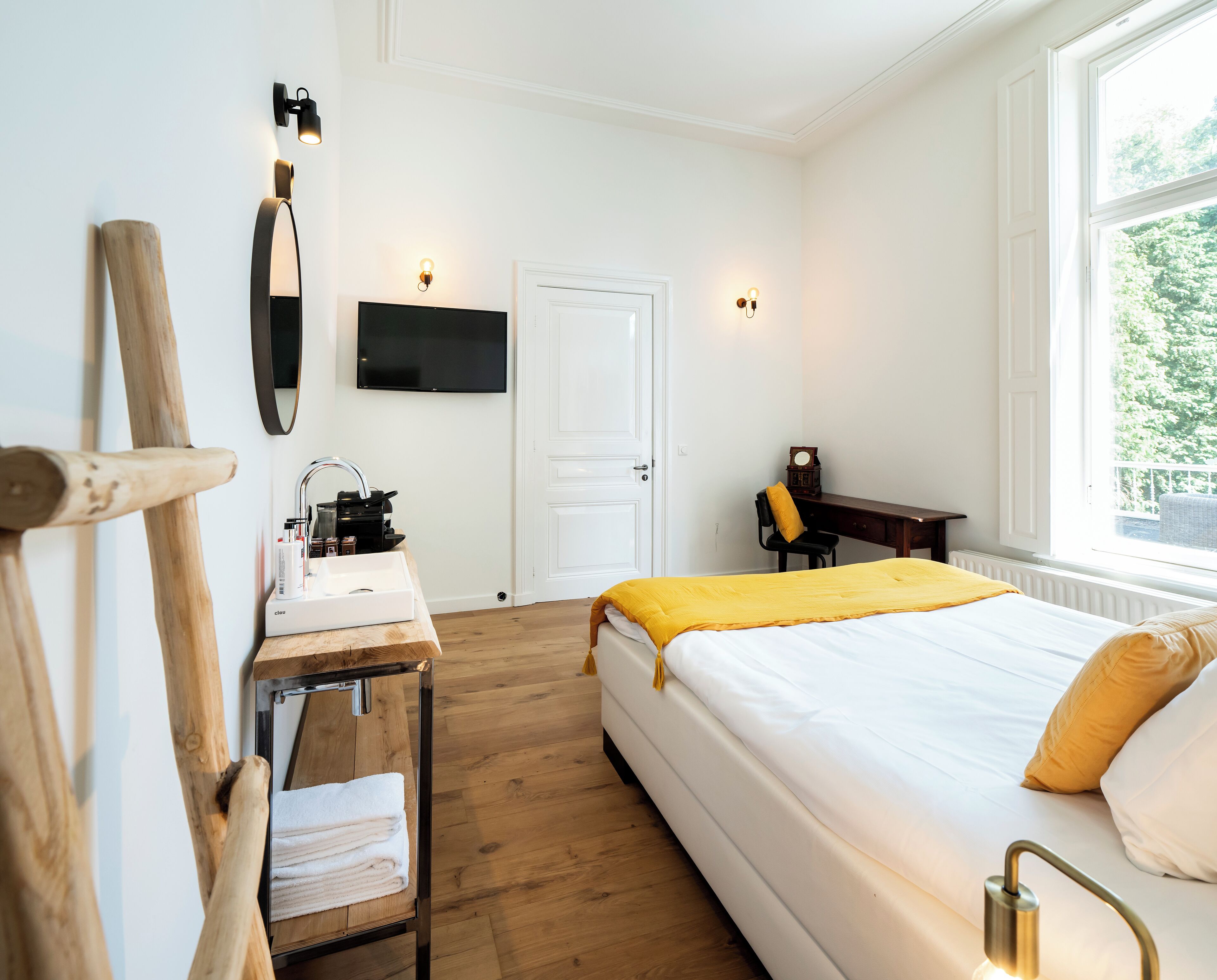 A hotel room with a bed, a desk with a sink, a chair, a TV mounted on the wall, and a window with a view of greenery.