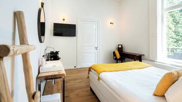 A hotel room with a bed, a desk with a sink, a chair, a TV mounted on the wall, and a window with a view of greenery.
