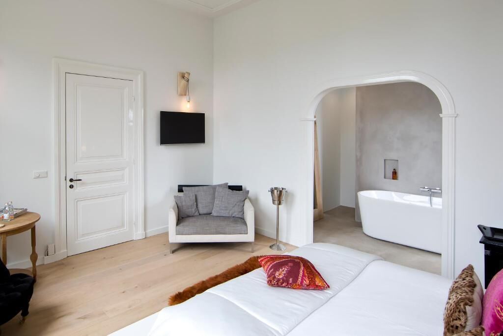 A bedroom with a bed, a grey armchair, a small table, a floor lamp, and a bathroom with a bathtub.
