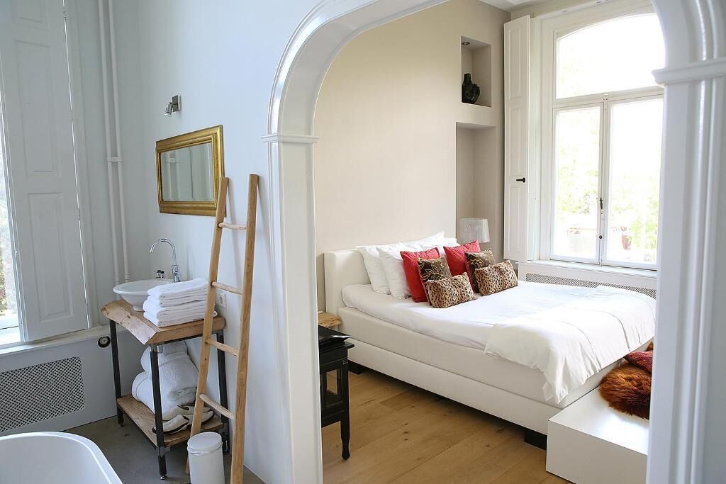 A bedroom with a white bed, a white dresser, a wooden ladder, a white radiator, and a window with a view of greenery.