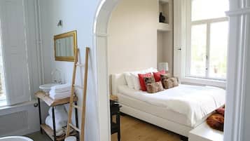 A bedroom with a white bed, a white dresser, a wooden ladder, a white radiator, and a window with a view of greenery.