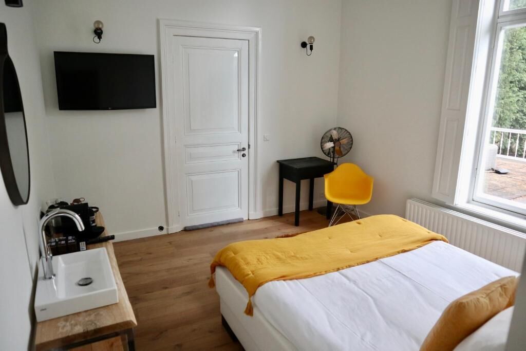 A bedroom with a bed, a bedside table with a sink, a TV mounted on the wall, a door, and a window.