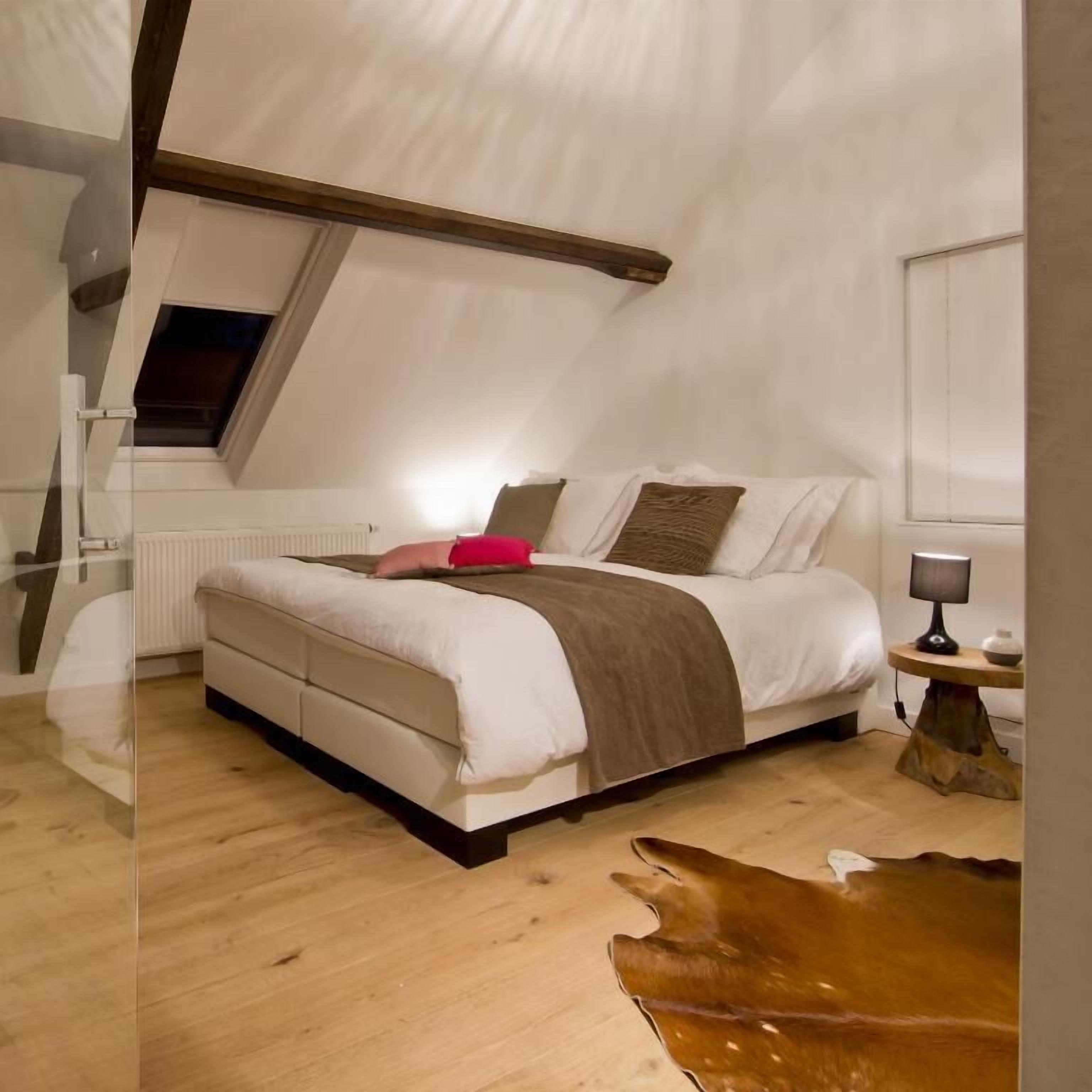 Un dormitorio con techo inclinado, cama con colcha blanca y marrón, almohada roja y suelo de madera.