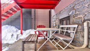 A covered outdoor seating area with a wooden table and two Adirondack chairs, a stone wall, and a red metal staircase. - Fox Hotel Palais Des Congres (Montréal)