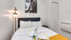 Een net opgemaakt bed met witte lakens, een nachtkastje, een aan de muur hangend kunstwerk, een vloerlamp en een witte deur.